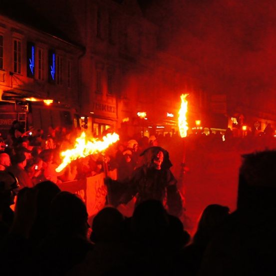 Eine Menschenmenge versammelt sich um eine Person mit Maske, die eine brennende Fackel hält. Die Szene spielt bei Nacht mit einem roten Schein. Mehrere Menschen schauen zu, einige halten ihre Telefone. Im Hintergrund befinden sich Gebäude mit beleuchteten Schildern.