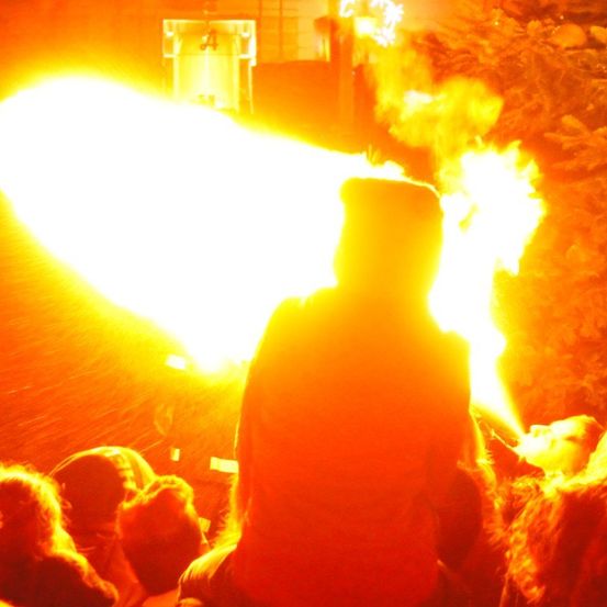 Eine Menschenmenge beobachtet, wie eine Person Feuer ausspuckt, während ein Feuerwehrmann hinter ihr steht, alles unter einem beleuchteten Schild und in der Nähe eines Baumes.