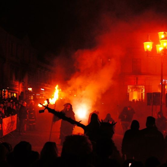Eine Menschenmenge versammelt sich nachts um ein großes Lagerfeuer, mit Menschen in Kostümen und mit Fackeln. Die Szene ist von roten Lichtern beleuchtet.