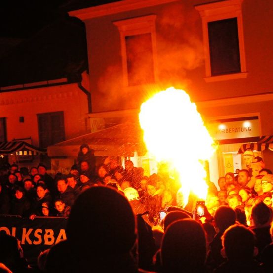 Eine Menschenmenge versammelt sich um ein großes Feuer, möglicherweise bei einem Festival, mit Gebäuden im Hintergrund und einem Banner sichtbar.