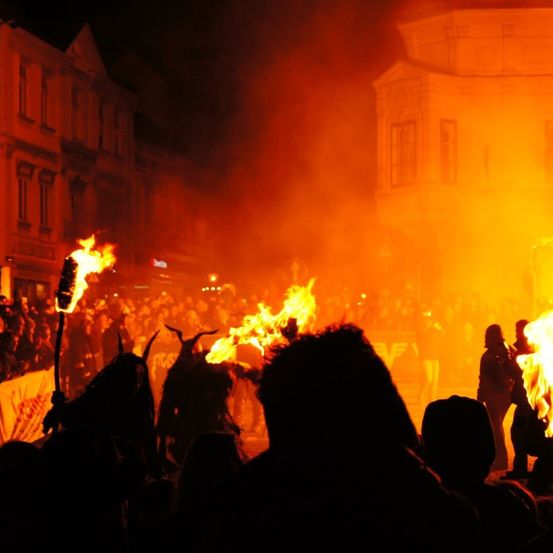Eine Menschenmenge ist um ein Lagerfeuer in der Nacht versammelt. Einige Personen tragen Hörnerkostüme. Das Feuer beleuchtet den Bereich und wirft Schatten auf die Gebäude.