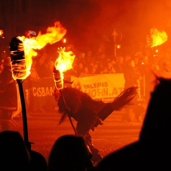 Ein Darsteller mit brennenden Fackeln unterhält eine Menge, silhouettiert vor einem Hintergrund aus Feuer und Rauch.