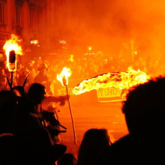 Eine Gruppe von Menschen auf einem öffentlichen Platz, die brennende Fackeln halten, wobei die Flammen ein oranges Licht auf die Menge und die Gebäude werfen.