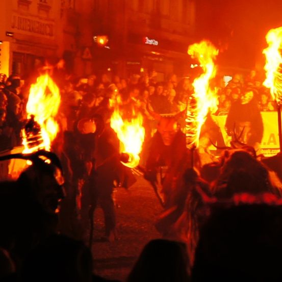 Eine Menschenmenge versammelt sich bei Nacht, um Darsteller mit Feuerfackeln zu beobachten. Einige Personen tragen Masken und halten Banner hoch.