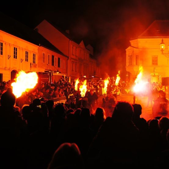 Eine nächtliche Veranstaltung mit einer Menschenmenge, die sich um Lagerfeuer versammelt. Die Gebäude im Hintergrund sind beleuchtet und werfen ein warmes Licht auf die Szene.