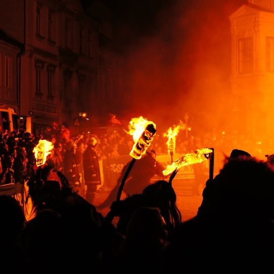 Eine Menschenmenge versammelt sich bei Nacht im Freien, wahrscheinlich für ein Festival, mit feuerbeleuchteten Fackeln und einer hellen orangefarbenen Flamme im Hintergrund.