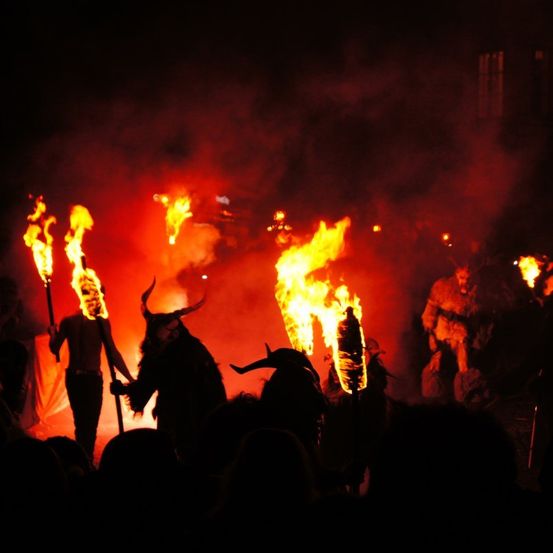 Menschen in Hörnermasken halten brennende Fackeln vor einem großen Feuer bei Nacht.