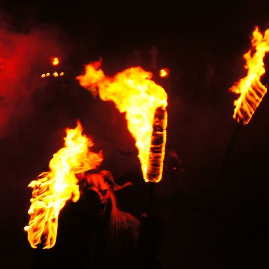 Drei brennende Fackeln beleuchten den dunklen Bereich, wobei die mittlere die intensivste Helligkeit ausstrahlt. Die Flammen sind leuchtend gelb, und die Umgebung ist dunkel.