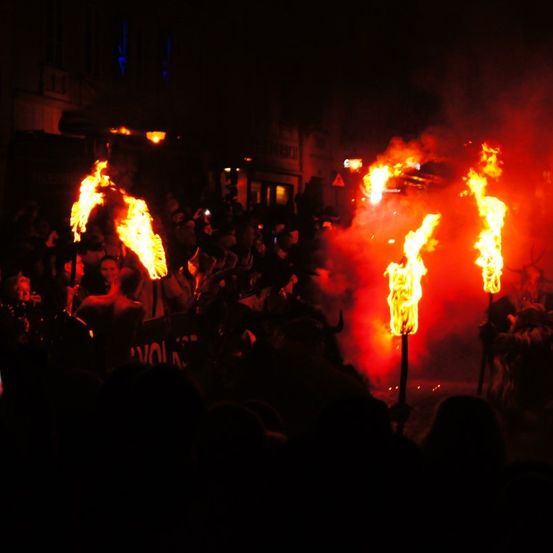 Eine Menschenmenge versammelt sich bei Nacht, erhellt von Fackeln mit Flammen. Menschen tragen Kostüme und einige halten Schilder hoch, was eine lebhafte Atmosphäre erzeugt.