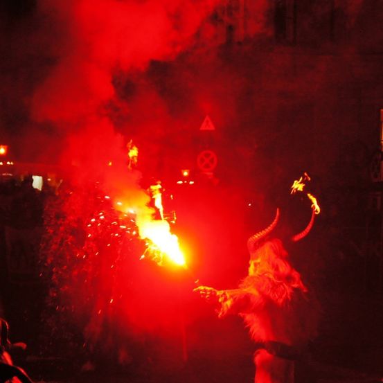Eine Person in einem gehörnten Kostüm, die eine brennende Fackel hält, umgeben von rotem Rauch. Die Szene spielt bei Nacht mit einem zuschauenden Publikum.