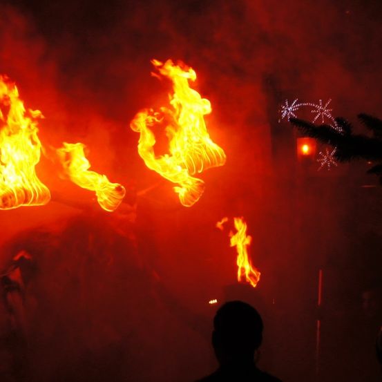 Eine Person steht vor einem großen Feuer, möglicherweise einem Lagerfeuer, mit Rauch und Schneeflocken im Hintergrund.