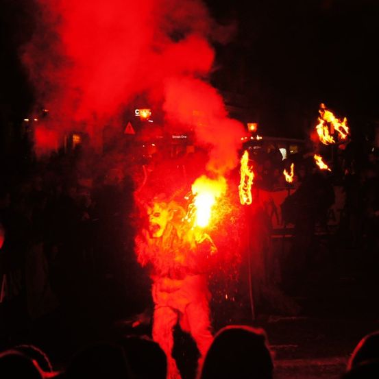Ein Künstler in einem Kostüm steht vor einer Menge, hält einen Feuerstab, bläst Feuer und ist von Rauch umgeben.