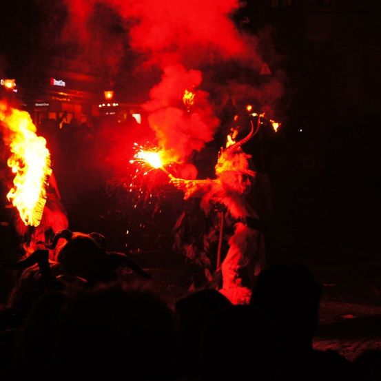 Eine Person in einem gehörnten Kostüm hält eine Fackel, die Funken sprüht, vor einer Menge, wobei eine andere Person eine Fackel mit Feuer hinter sich hält.