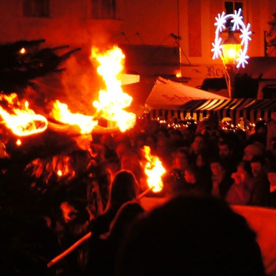 Ein Lagerfeuer brennt hell in der Nacht, mit einer Menschenmenge, die sich darum versammelt hat. Es gibt auch Lichter und Dekorationen in der Umgebung.