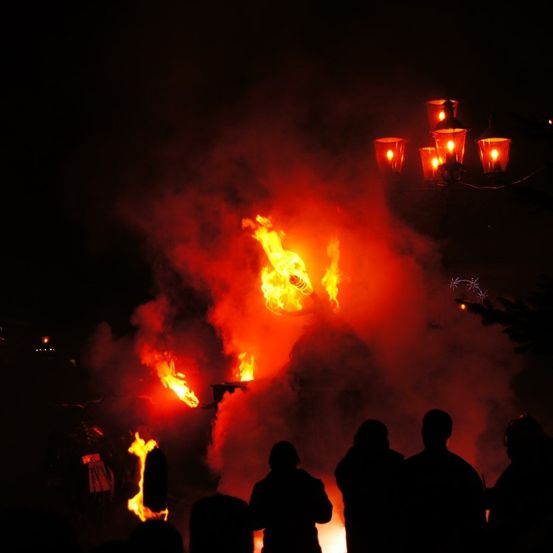 Eine Menschenmenge versammelt sich bei Nacht um ein Lagerfeuer, bei dem ein Feuerkunstwerk hell brennt und Schatten auf die Gesichter der Zuschauer wirft.