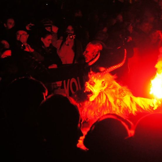Eine Menschenmenge beobachtet einen Darsteller in einem Löwenkostüm mit feuerroter Mähne und gehörntem Masken Gesicht, der auf der Bühne tanzt und brüllt.