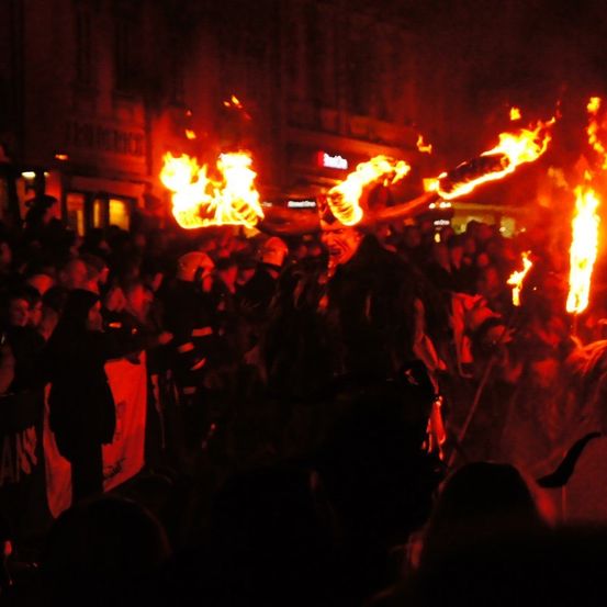 Ein nächtliches Festival mit einer Menschenmenge, die einen Darsteller mit brennenden Fackeln umgibt. Der Darsteller scheint zu schreien oder zu singen, während er von Flammen umgeben ist.