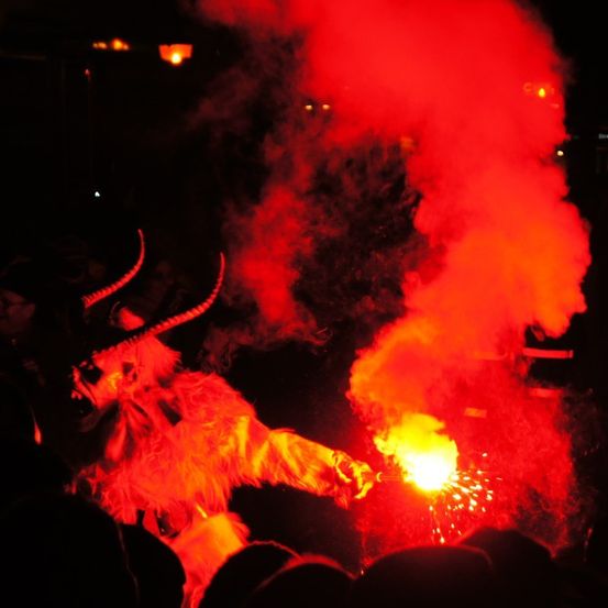 Eine Figur in einer gehörnten Maske und Fellanzug steht unter rotem Rauch und Funken und tritt bei einer nächtlichen Veranstaltung auf.