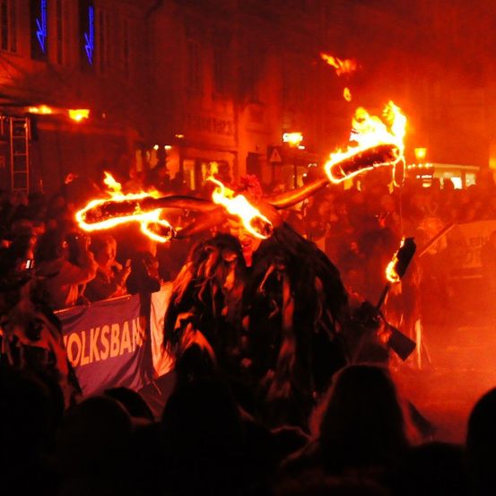 Ein Performer in einem gehörnten Kostüm mit brennenden Fackeln steht inmitten einer Menge während einer nächtlichen Veranstaltung.