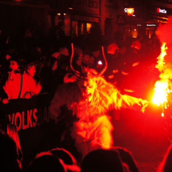 Eine Menschenmenge bei Nacht mit einer Person in einem Wolfskostüm, die ein Feuer hält. Im Hintergrund hält eine Person eine Kamera und ein Banner sagt 'VOLKS.AN'.