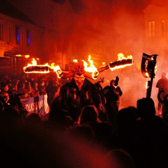 Eine maskierte Figur mit brennenden Fackeln steht mitten in einer Menschenmenge bei Nacht, mit Gebäuden und Rauch im Hintergrund.