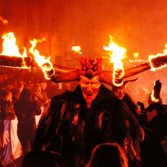 Eine Person in einem dämonischen Kostüm mit brennenden Fackeln an den Armen wird von einer Menge umringt.