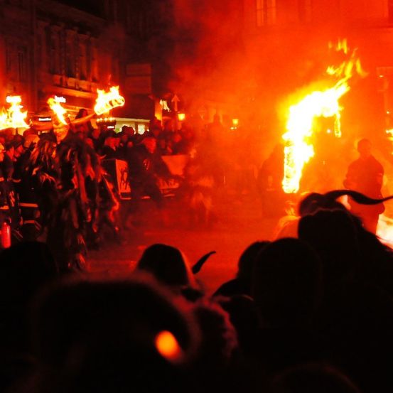 Eine nächtliche Versammlung mit Menschen, die brennende Fackeln halten, die ein warmes Licht und Schatten werfen. Gebäude säumen den Hintergrund.