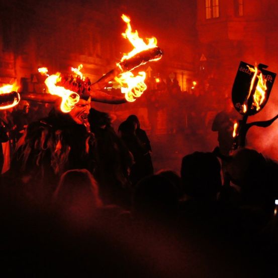 Eine Gruppe von Menschen versammelt sich bei Nacht und beobachtet einen Mann in einem feuerspuckenden Kostüm mit brennenden Hörnern und Flammen. Die Szene spielt sich in einem dunklen Bereich ab, mit verschwommenen Figuren im Hintergrund.