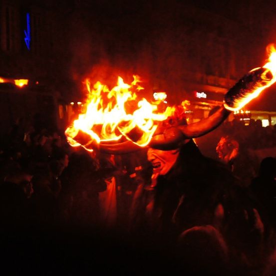 Eine als Teufel verkleidete Person hält einen brennenden Horn in einer nächtlichen Feier mit einer Menge um sie herum.