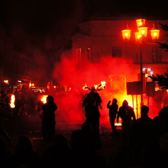 Eine Gruppe von Menschen in Kostümen steht bei Nacht vor einem Feuer. Das Feuer produziert roten Rauch. Andere Menschen stehen vor einem Gebäude.