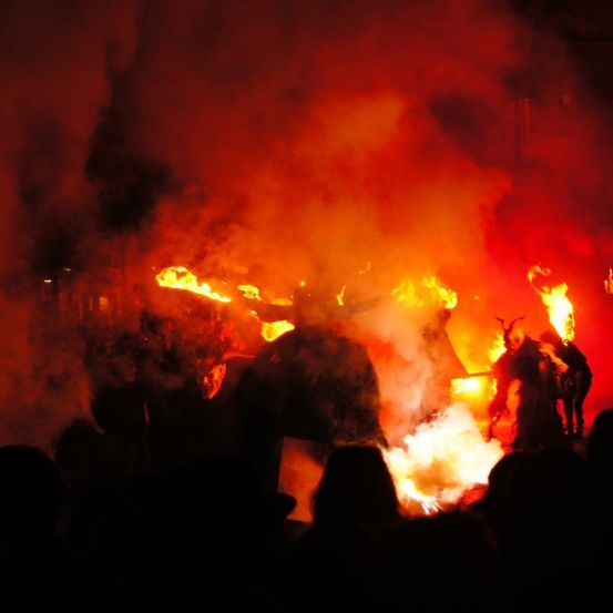 Eine Menge versammelt sich um ein Lagerfeuer mit Menschen in Kostümen und Masken. Das Feuer brennt hell und ist von Rauch umgeben.