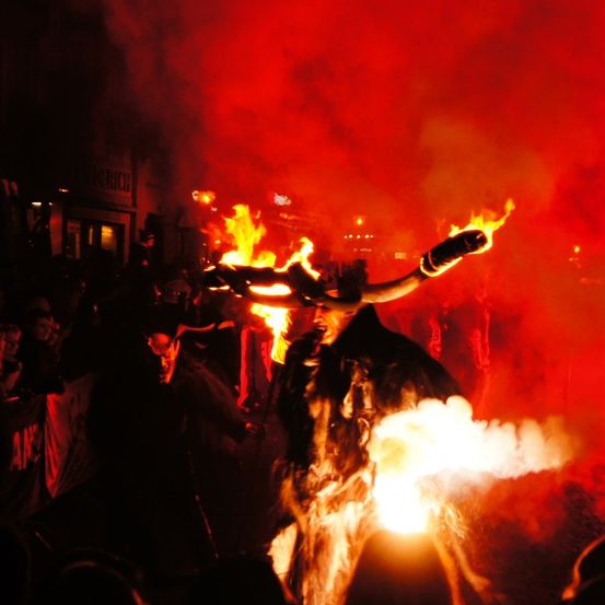 Eine Person in einer feuerspeienden Maske führt einen Ritualakt in einem dunklen Umfeld durch. Der Hintergrund ist mit rotem Rauch und Flammen gefüllt.