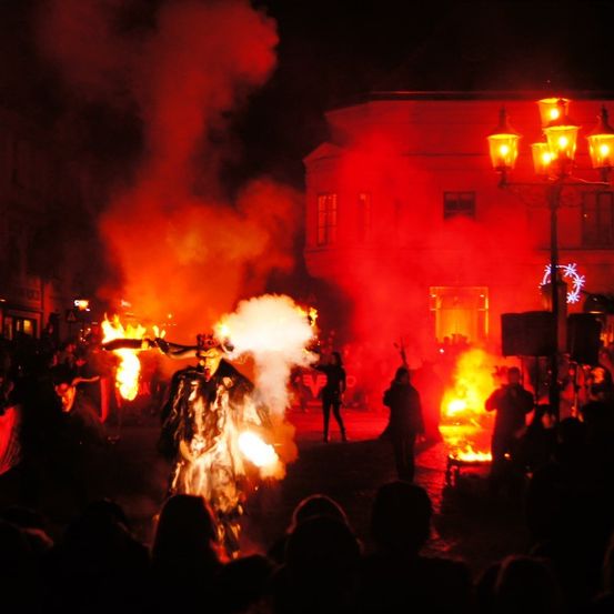 Eine Menschenmenge versammelt sich um ein Feuer, bei dem ein Künstler eine Fackel hält. Das Feuer ist von Rauch umgeben, und die Menge wird vom Licht des Feuers beleuchtet.