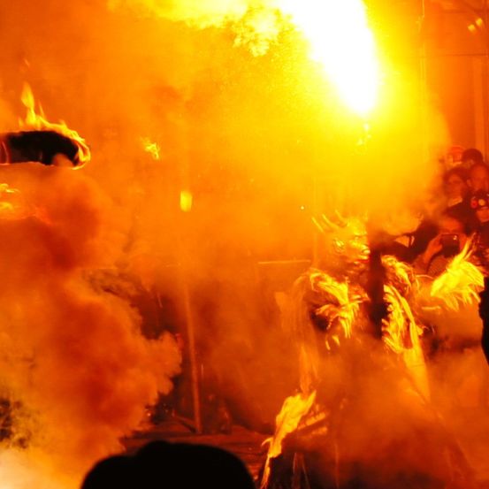 Ein Feuer speiender Darsteller, umgeben von Rauch, mit einer Menge im Hintergrund, eine Person trägt einen Helm und hält eine Kamera.