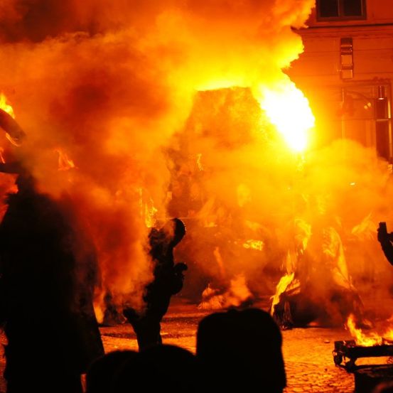 Eine nächtliche Szene mit einem brennenden Götzenbild, umgeben von Rauch, besucht von Menschen und Silhouetten im Vordergrund.