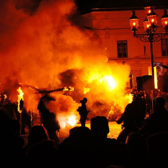 Ein nächtliches Festival mit einer großen Feuershow. Mehrere Menschen sind versammelt und beobachten, wie andere mit Feuer auftreten. Das Feuer beleuchtet die Gesichter der Menge.