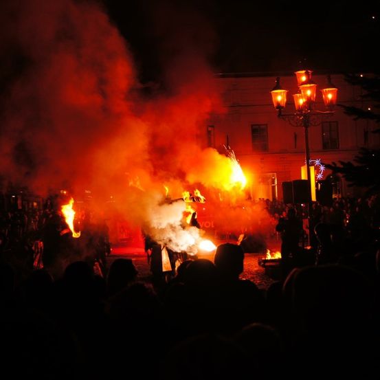 In der Nacht versammelt sich eine Menschenmenge um ein zentrales Lagerfeuer, wobei zwei Künstler inmitten der Flammen stehen. Ein Laternenpfahl mit blauen Lichtern und ein geschmückter Baum sind im Hintergrund zu sehen.