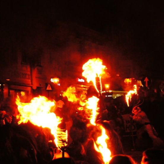 Ein nächtliches Festival zeigt große, leuchtende Flammen, wobei Menschen Fackeln halten und andere von unten zusehen, in einem schwach beleuchteten Bereich.