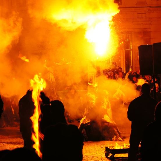 Eine Menschenmenge versammelt sich um ein Lagerfeuer bei Nacht, mit einer großen Flamme, die aufsteigt, und Leuten, die Fotos machen. Ein Lautsprecher steht rechts.