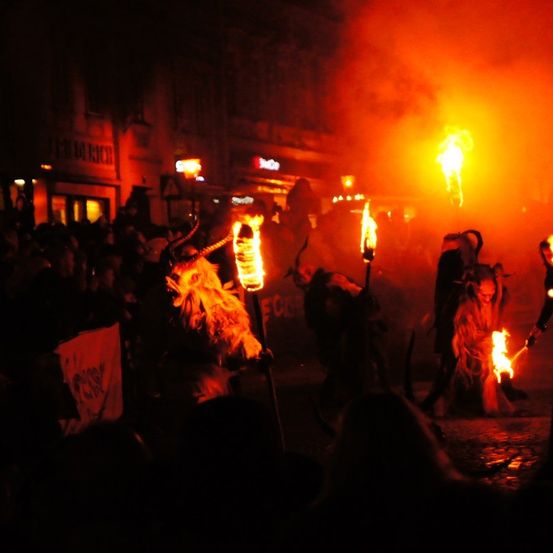 Eine Menschenmenge versammelt sich bei Nacht, wobei Personen in Kostümen brennende Fackeln halten. Sie stehen vor einem Gebäude mit einem Schild und im Hintergrund ist ein beleuchtetes Gebäude.