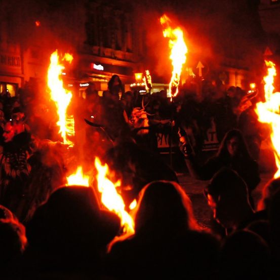 Ein nächtliches Festival mit einer Menschenmenge um ein großes Lagerfeuer, mehrere Personen halten Fackeln und tragen Kostüme.