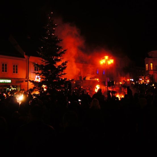 Eine Menschenmenge versammelt sich nachts um einen beleuchteten Weihnachtsbaum und mehrere Lagerfeuer. Gebäude mit beleuchteten Fenstern umgeben die Szene.