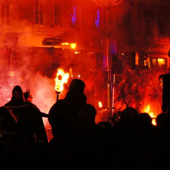 Eine Menschenmenge versammelt sich um ein Lagerfeuer in einem schwach beleuchteten Bereich, mit aufsteigenden Flammen und Rauch. Eine Person hält eine Fackel, und eine andere trägt einen Helm. Ein Gebäude ist im Hintergrund zu sehen.