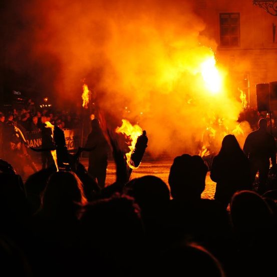 Eine Menschenmenge versammelt sich bei Nacht für ein Lagerfeuer-Event, einige halten brennende Fackeln und andere schauen zu. Rauch und Flammen steigen im Vordergrund auf.