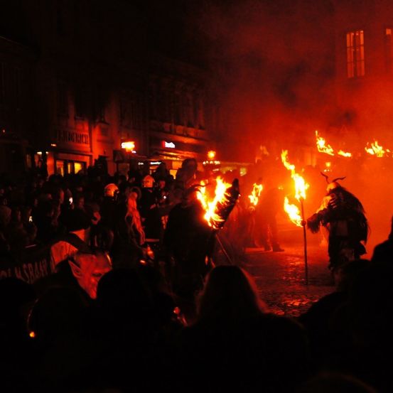 Ein nächtliches Festival mit Menschen, die Fackeln halten und Hörnermasken tragen. Eine Menge beobachtet von der Straße.