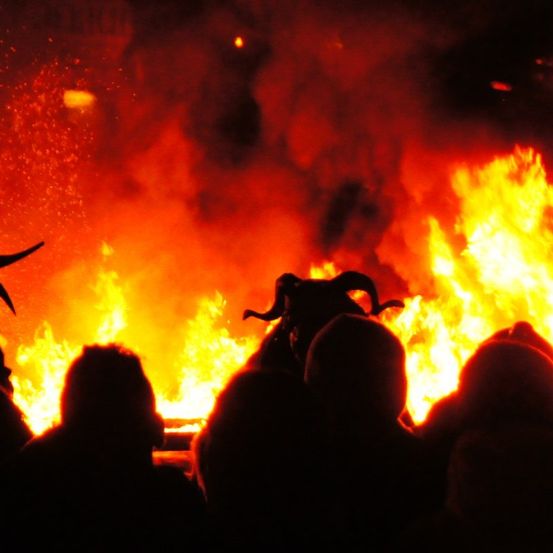 Ein großes Lagerfeuer mit Flammen und Rauch, umgeben von Silhouetten von Menschen, möglicherweise ein Festival oder Ritual.