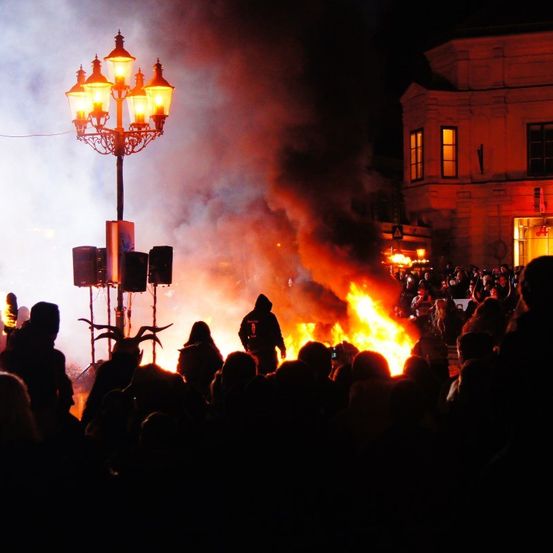 Eine Menschenmenge versammelt sich bei Nacht um ein Lagerfeuer, mit aufsteigendem Rauch und Flammen. Eine Person steht in der Nähe des Feuers, und eine Straßenlampe erhellt die Szene.