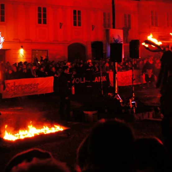 Eine große Menschenmenge versammelt sich um ein mit roten Lichtern beleuchtetes Feuer. Eine Person hält eine Fackel, und Schilder mit Bannern sind sichtbar.