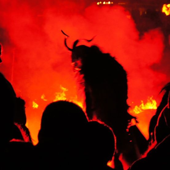Eine Gruppe von Menschen beobachtet einen Darsteller in einer gehörnten Maske, der vor einem großen Feuer tanzt.