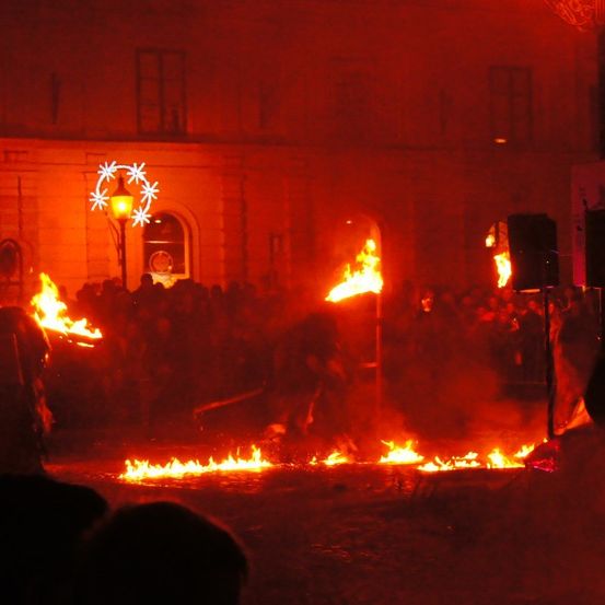 Eine Menschenmenge versammelt sich um eine Feuershow, die Flammen beleuchten ein Gebäude mit festlichen Dekorationen.